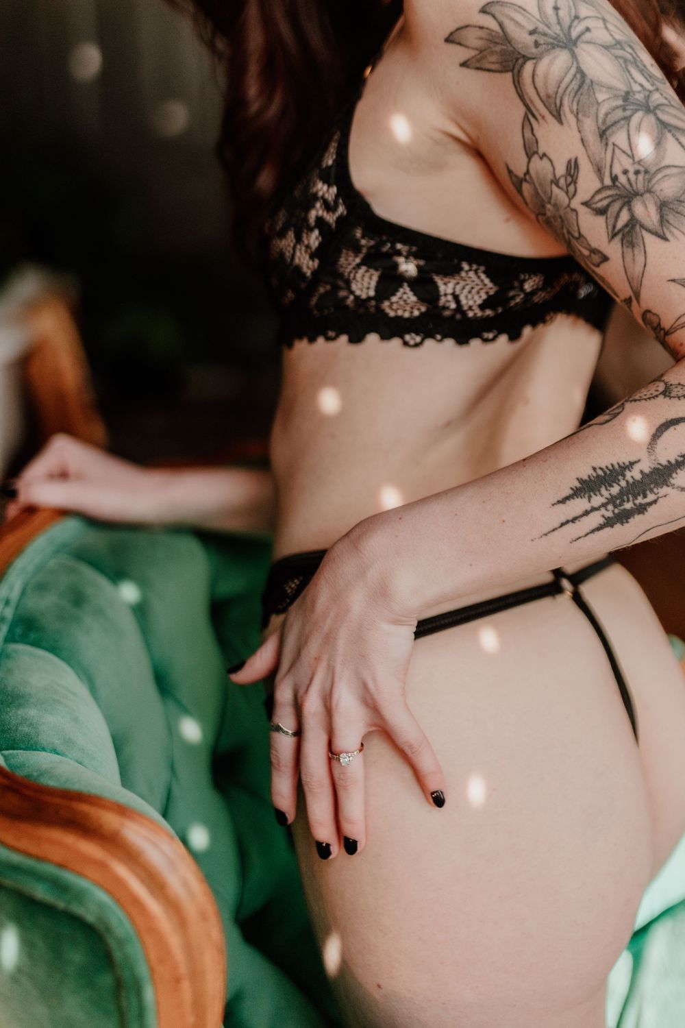 Woman in black lace lingerie with floral tattoos poses on green velvet chair.