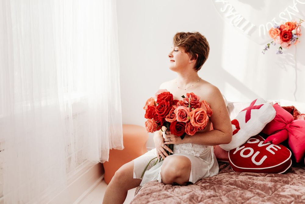 Young man sitting on a bed holding a bouquet of red and orange roses with Valentine's Day decorations nearby.