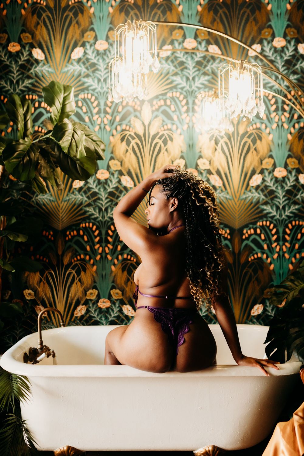 Woman in lingerie sitting in vintage clawfoot bathtub with art nouveau wallpaper and chandelier.