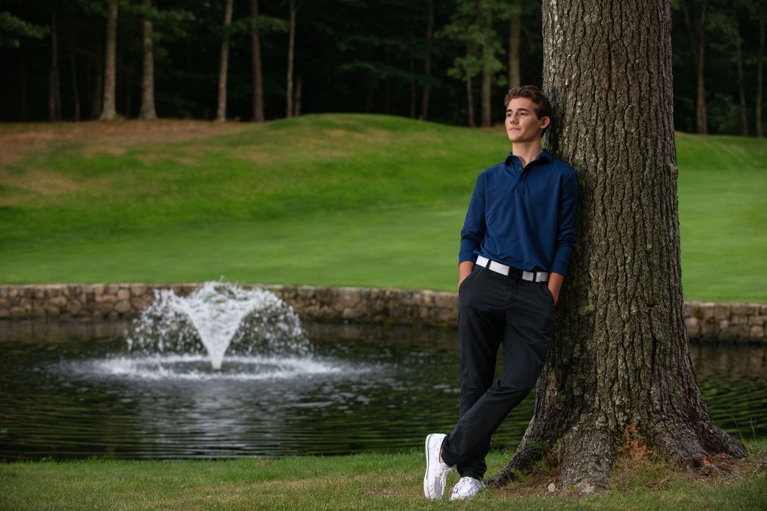 Person in navy jacket posing by large tree trunk with fountain in background on golf course.
