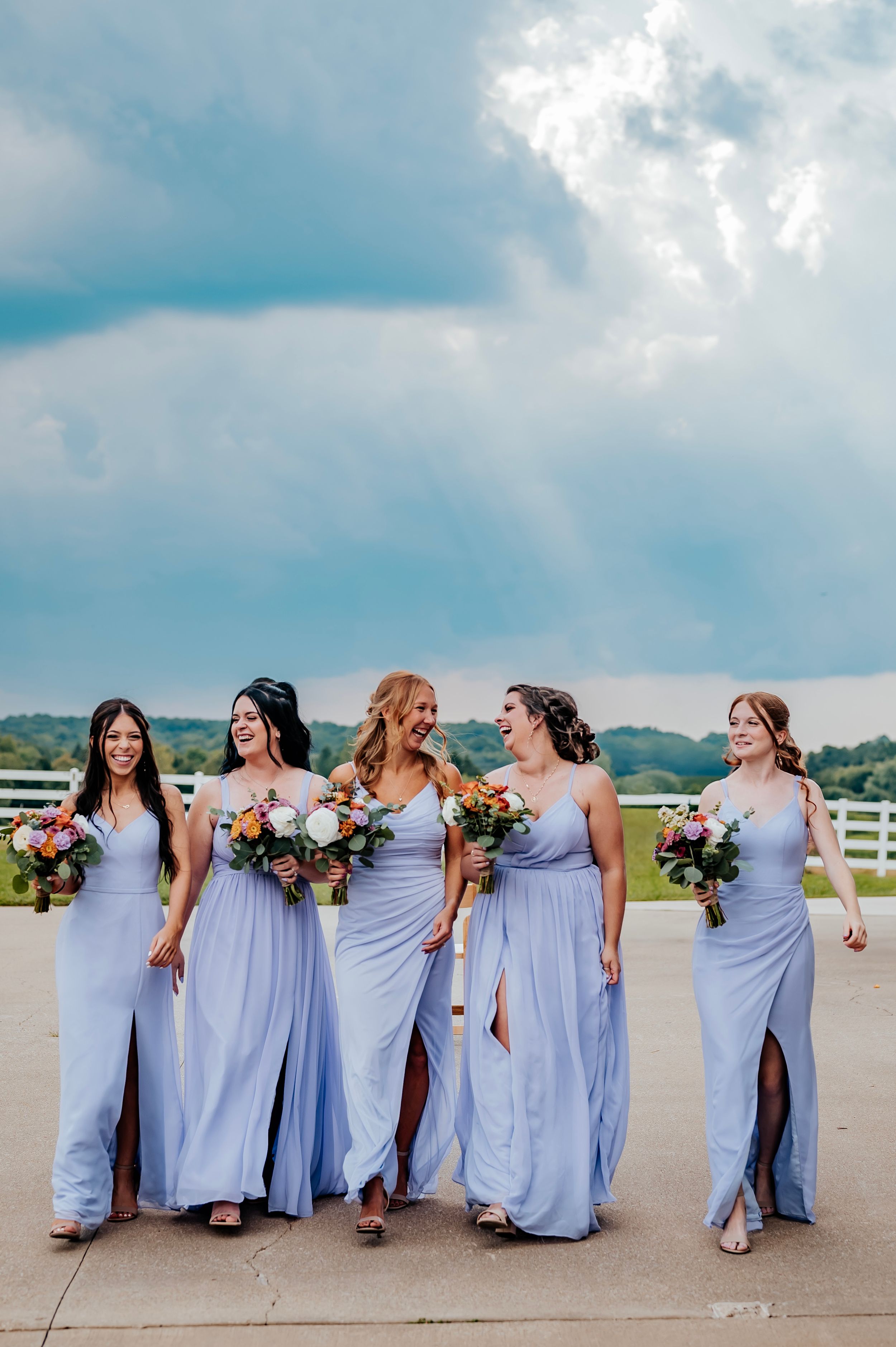 Bridesmaids in lavender dresses posing playfully against cloudy sky.