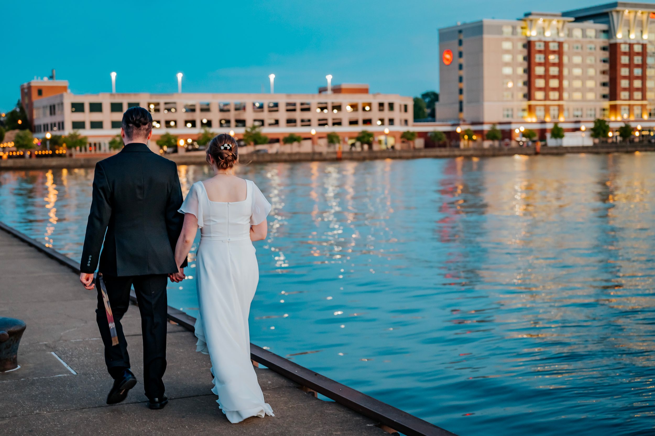 Couple walking along a waterfront promenade at sunset with city buildings in the background.