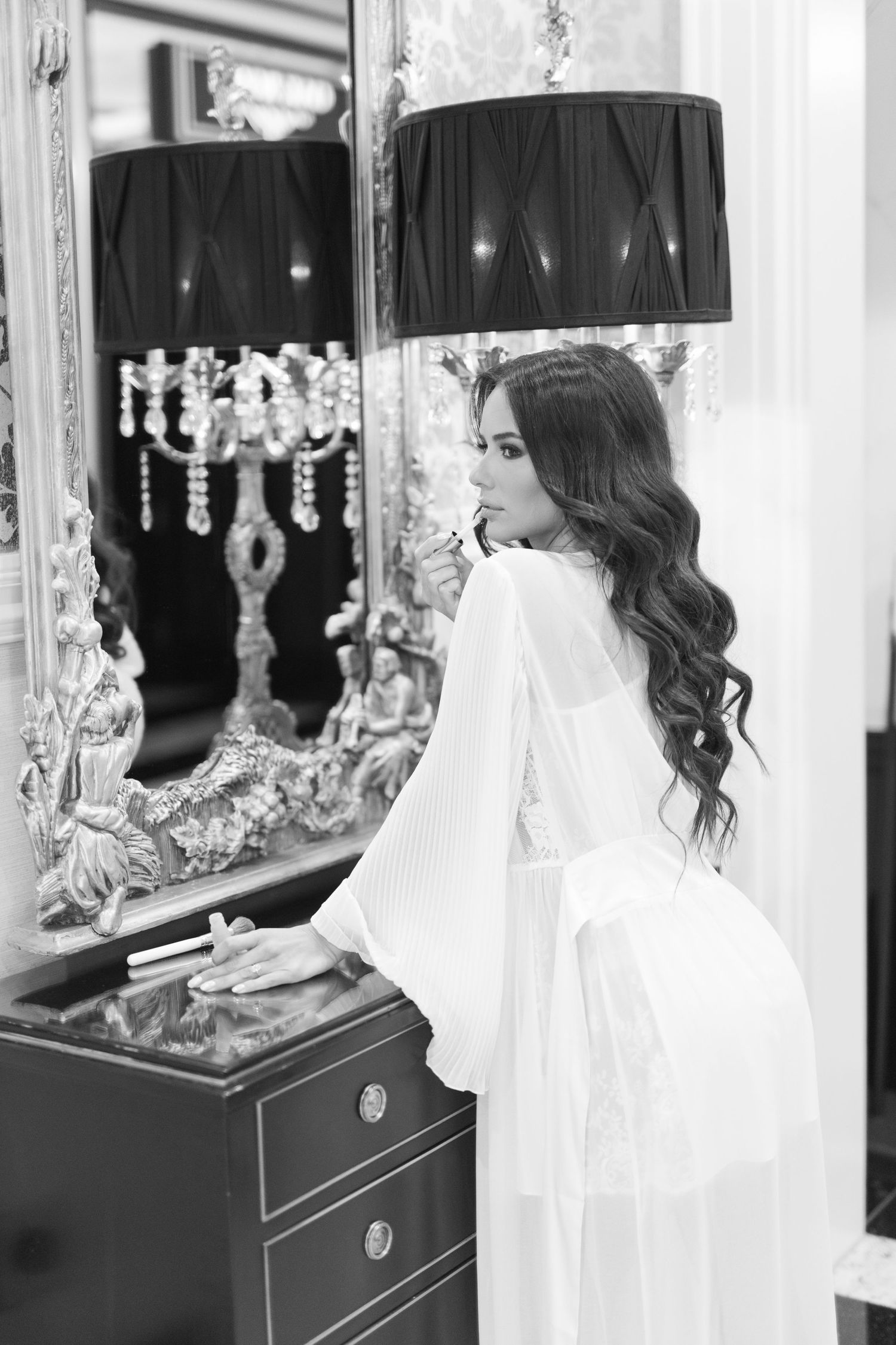 Black and white photo of bride in white robe applying lipstick before ornate gold mirror.