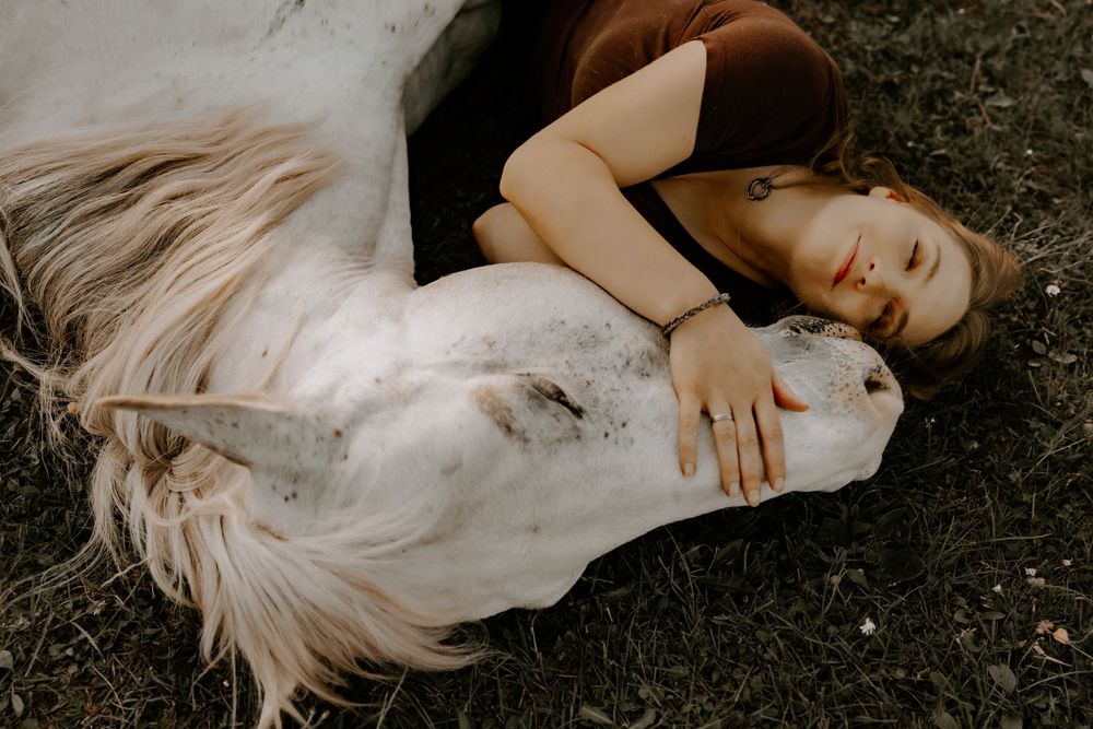 Person in black dress lying on grass while embracing a white horse outdoors.