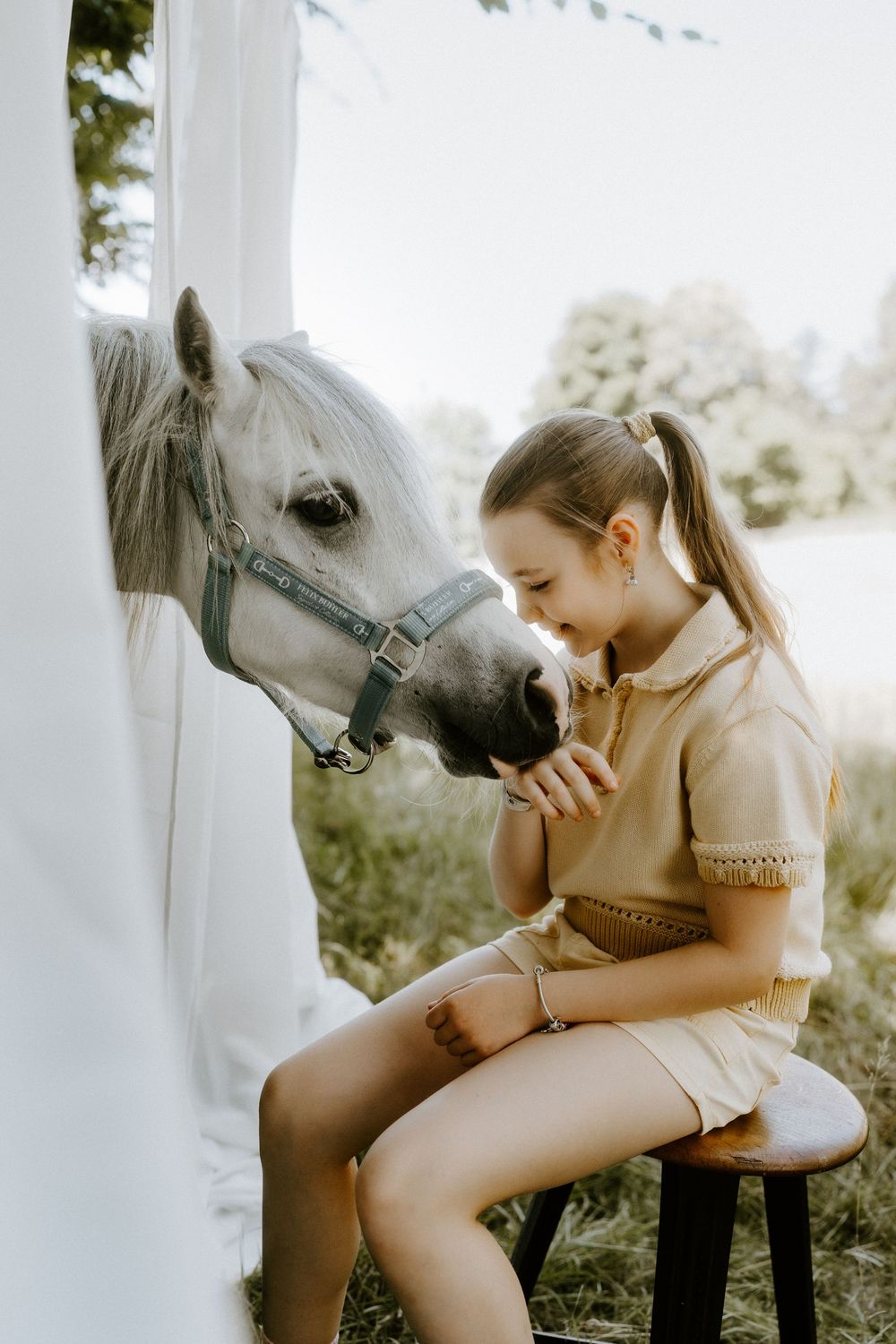 A young girl in a beige outfit sits on a stool, gently bonding with a white pony wearing a halter outdoors.