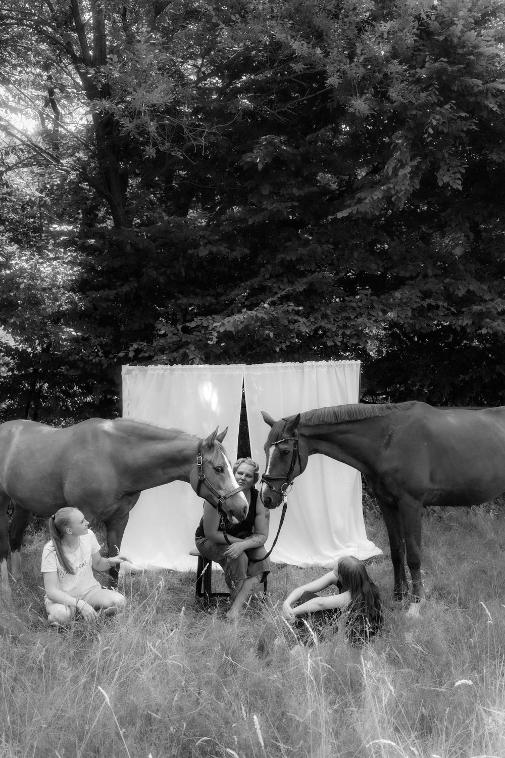 Black and white photo of two horses with a person and two girls sitting in a grassy field near white drapes and trees.