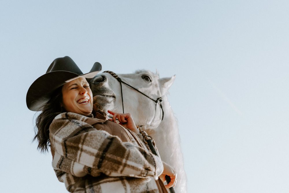 A joyful rider in a brown plaid blanket and cowboy hat smiles while sitting atop a white horse against a bright sky.