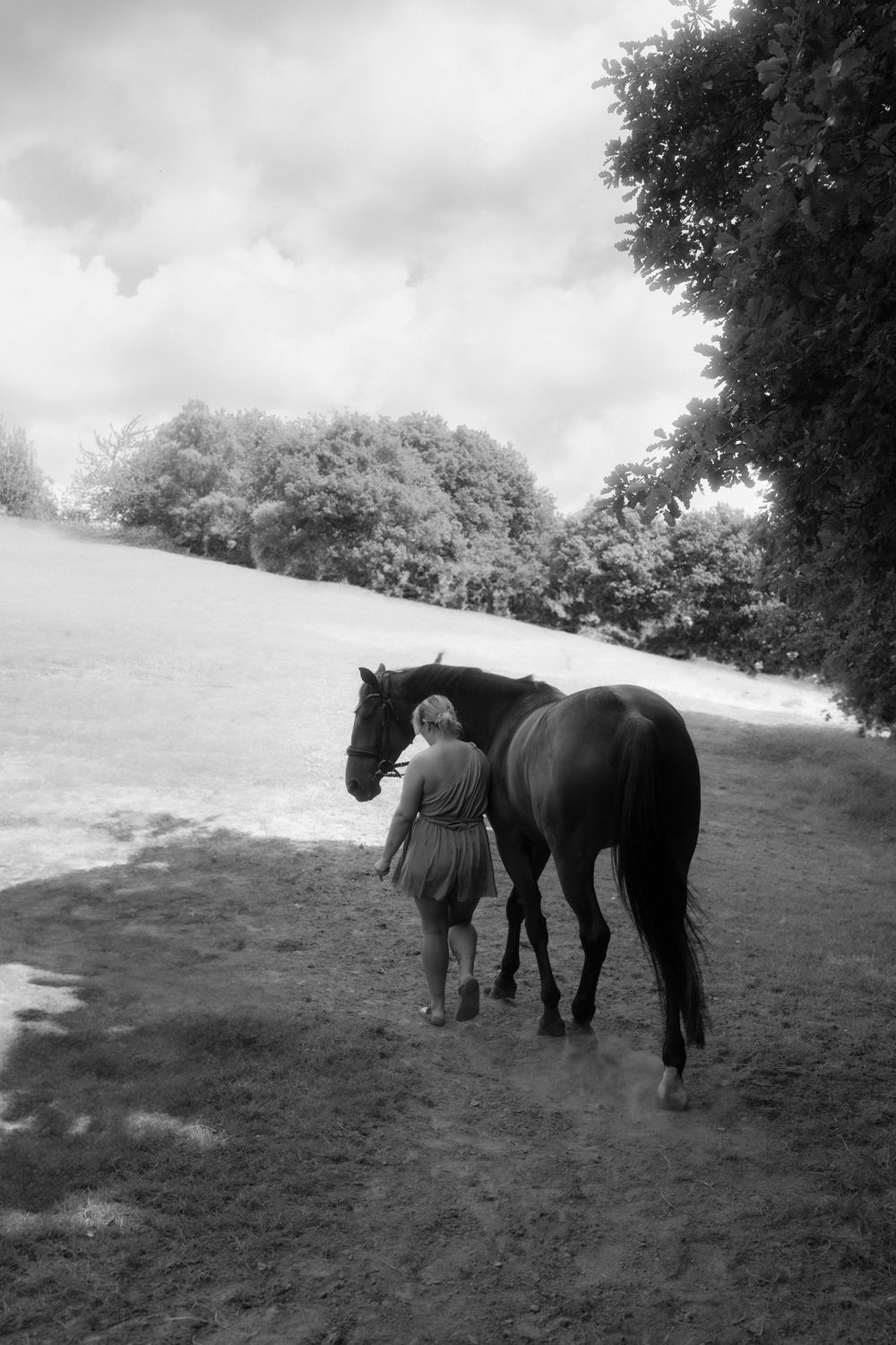 Black and white photo of a woman walking alongside a dark horse in a rural field with trees in the background.