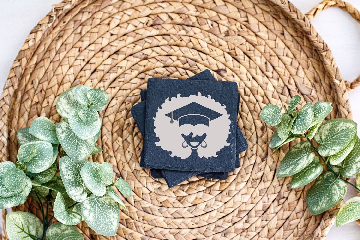 Graduation cap design on navy blue card centered on circular woven placemat with green leaves.