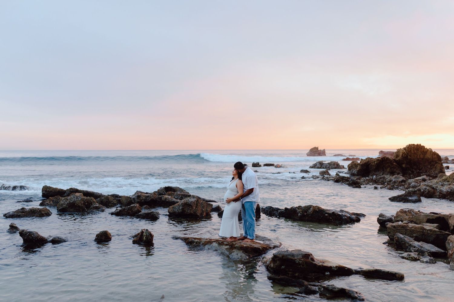 Two figures stand together on rocky coastal shore during sunset.