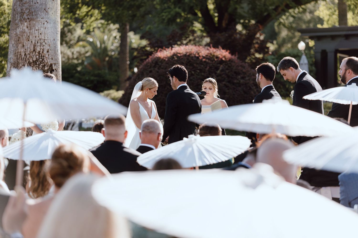 Outdoor wedding ceremony under white parasols surrounded by trees and natural lighting.