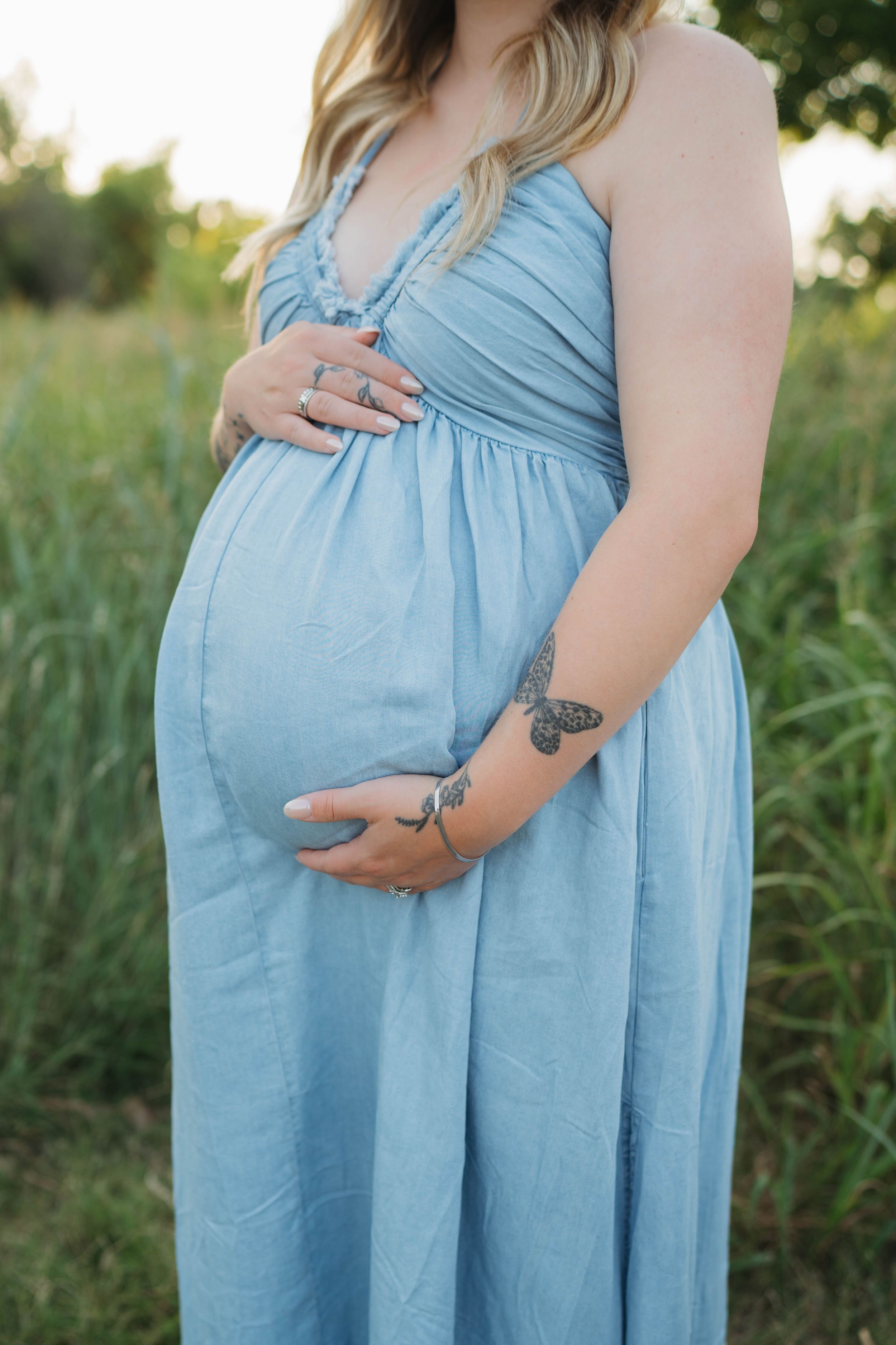 Close up of pregnant woman's belly in light blue maxi dress showing tattoo on arm.