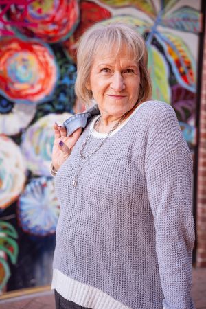 A person in a gray knit sweater poses against a colorful graffiti wall with a bright smile.