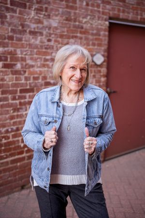 Senior woman in denim jacket and gray sweater poses playfully against brick wall background.