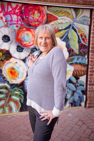 A person in a gray sweater smiles while posing in front of a vibrant floral wall mural.