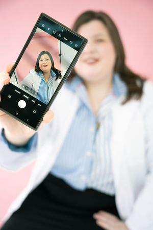 A smartphone displays a photo being taken against a pink background while showing a mirror reflection composition.