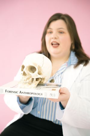 A person in a white lab coat and striped shirt holds and examines a human skull against a pink background.