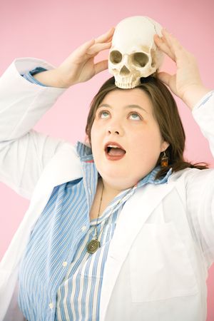 A person playfully balances a skull on their head while wearing a lab coat and striped shirt against a pink backdrop.