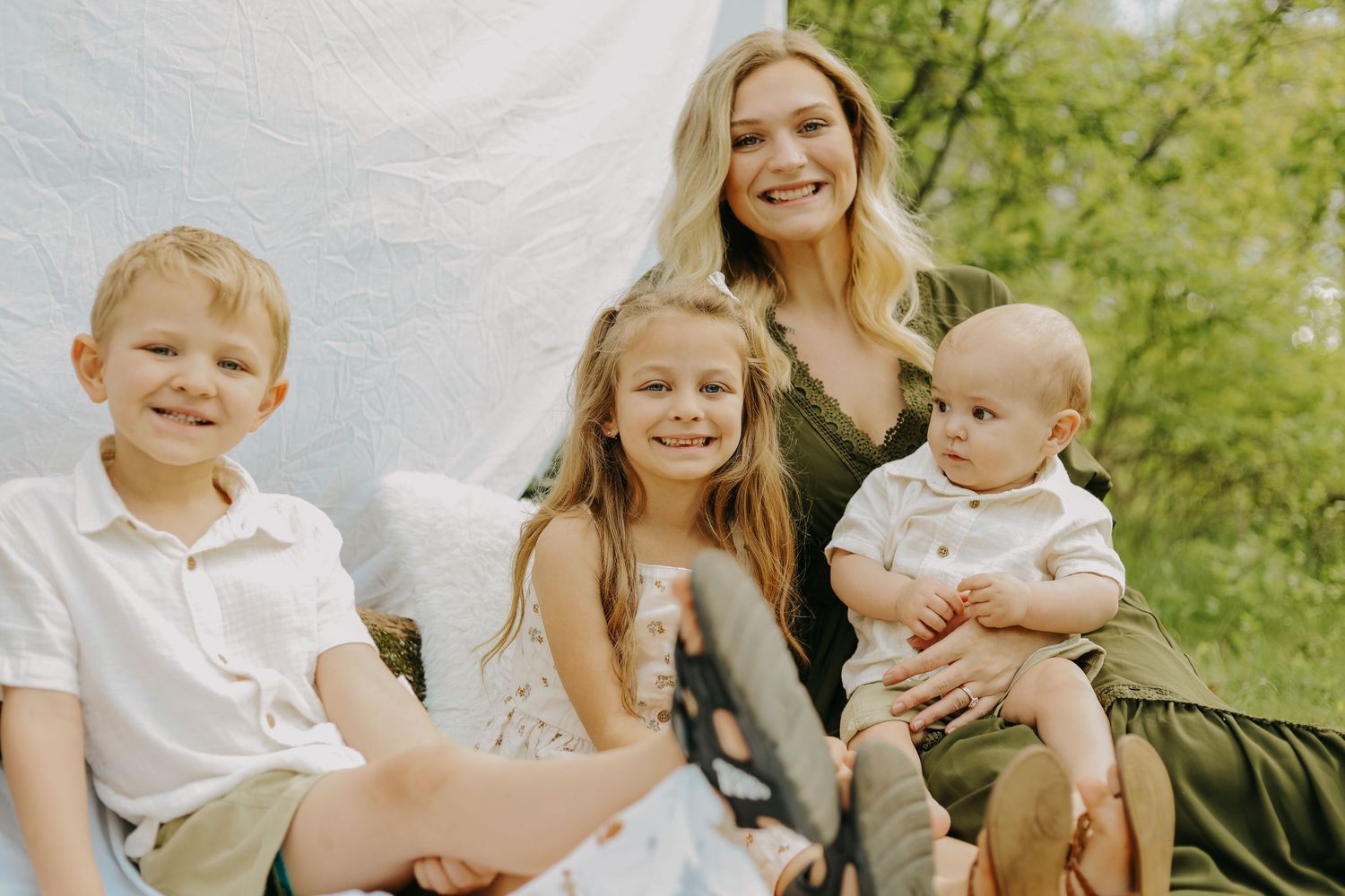 A family portrait outdoors against a white backdrop with children and a baby.