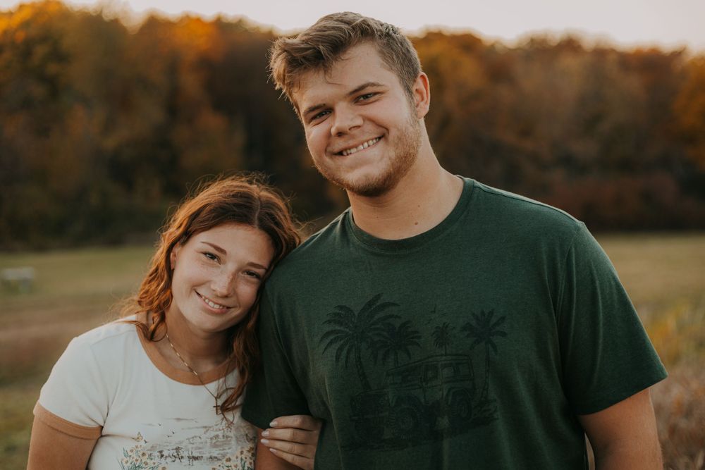 A couple smiles at the camera while embracing outdoors with fall foliage in the background.
