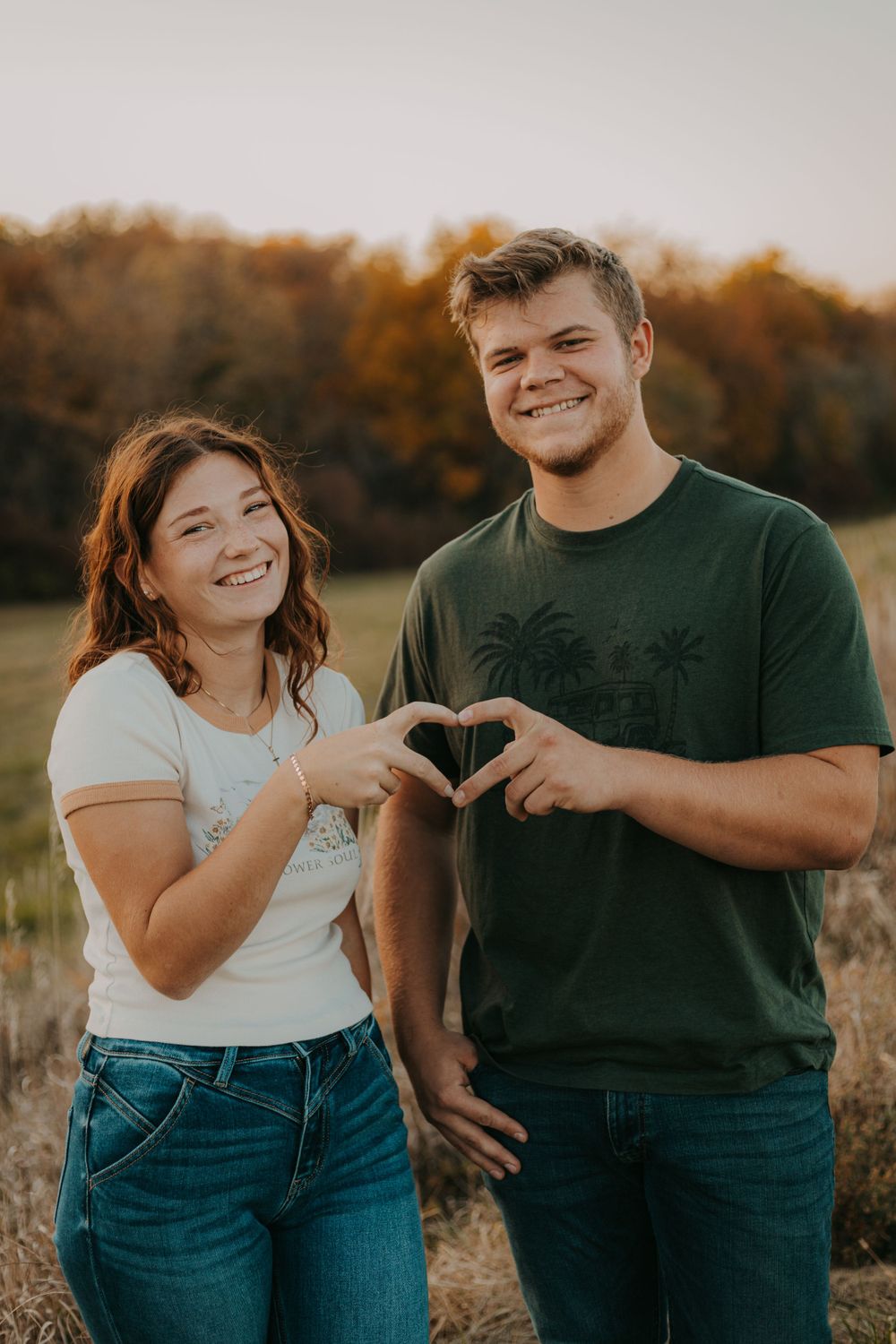 A couple makes a heart shape with their hands while posing together in a field at sunset.