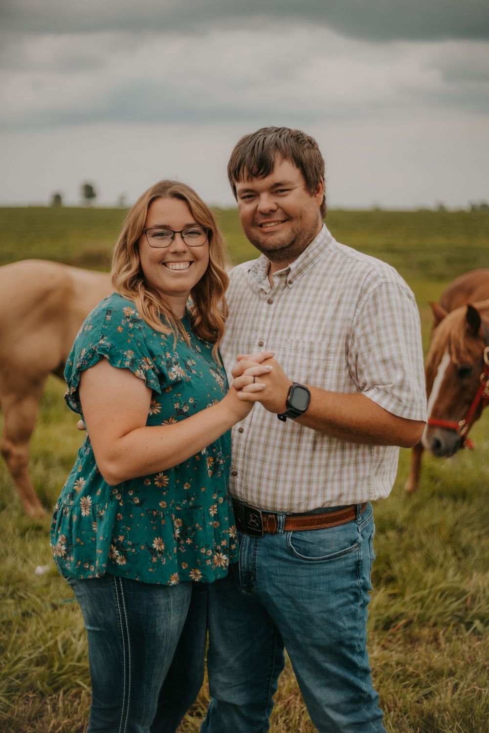 Couple poses together in green field with horses visible in background during engagement shoot.