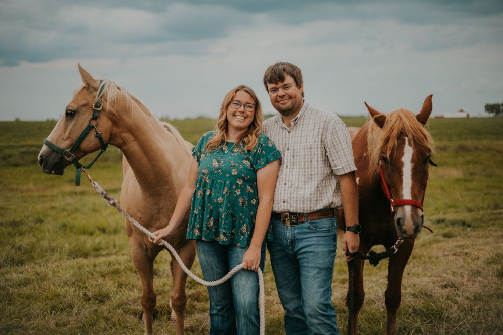 A couple stands between two horses in a rural field under overcast skies.