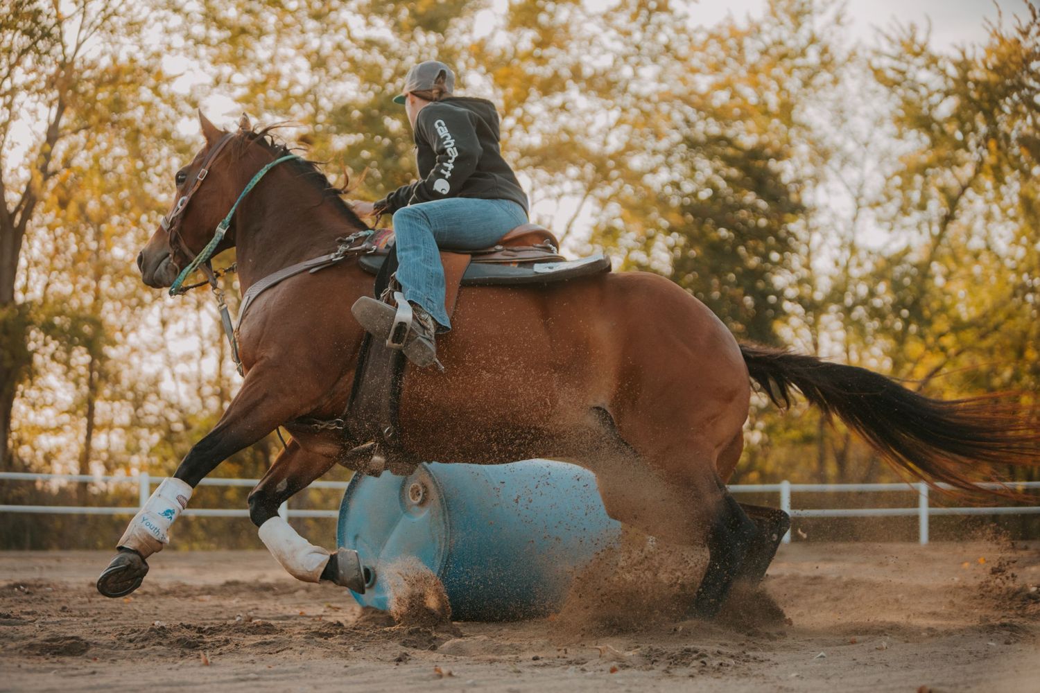 A rider in blue performs a barrel racing maneuver on a brown horse, kicking up arena dust.