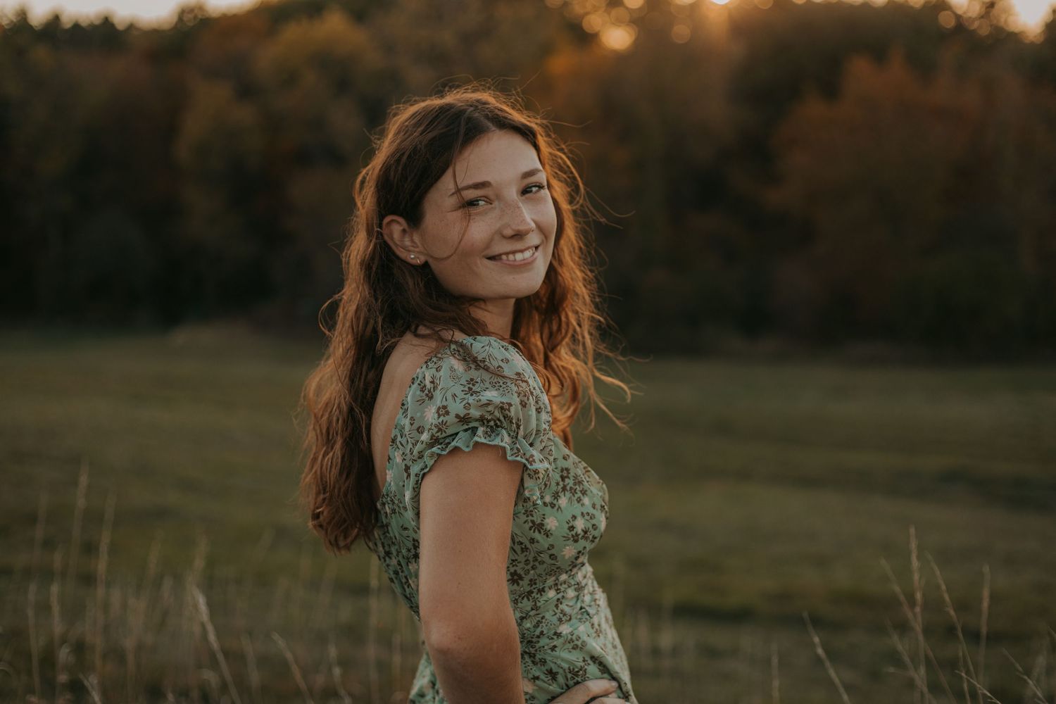 A young woman in a floral dress smiles over her shoulder in a golden field at sunset.