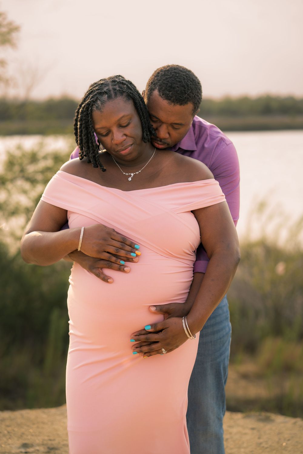 Intimate maternity photo of couple embracing on beach at sunset in pink dress and purple shirt.