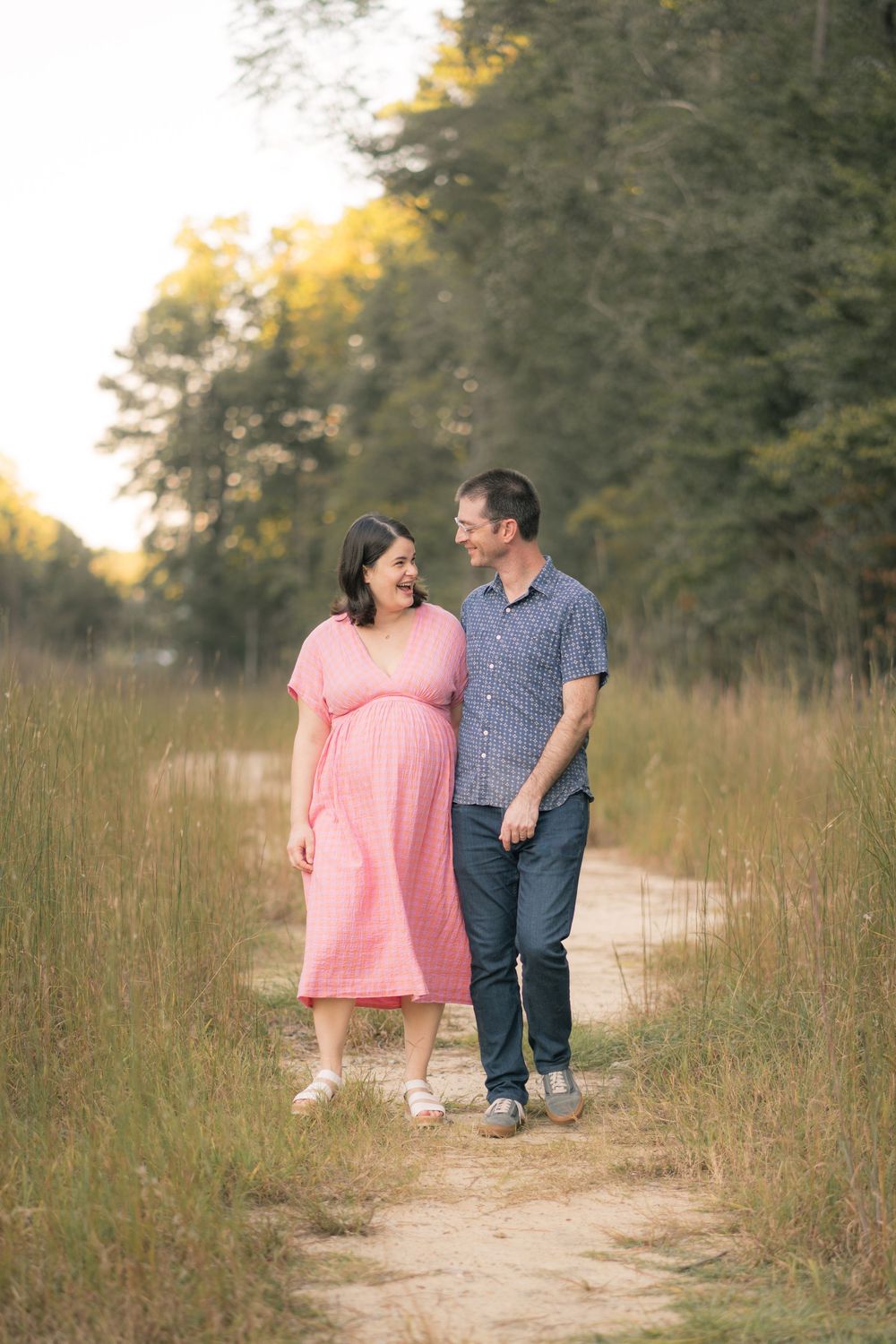 A couple walking together on a dirt path through tall grass at sunset.
