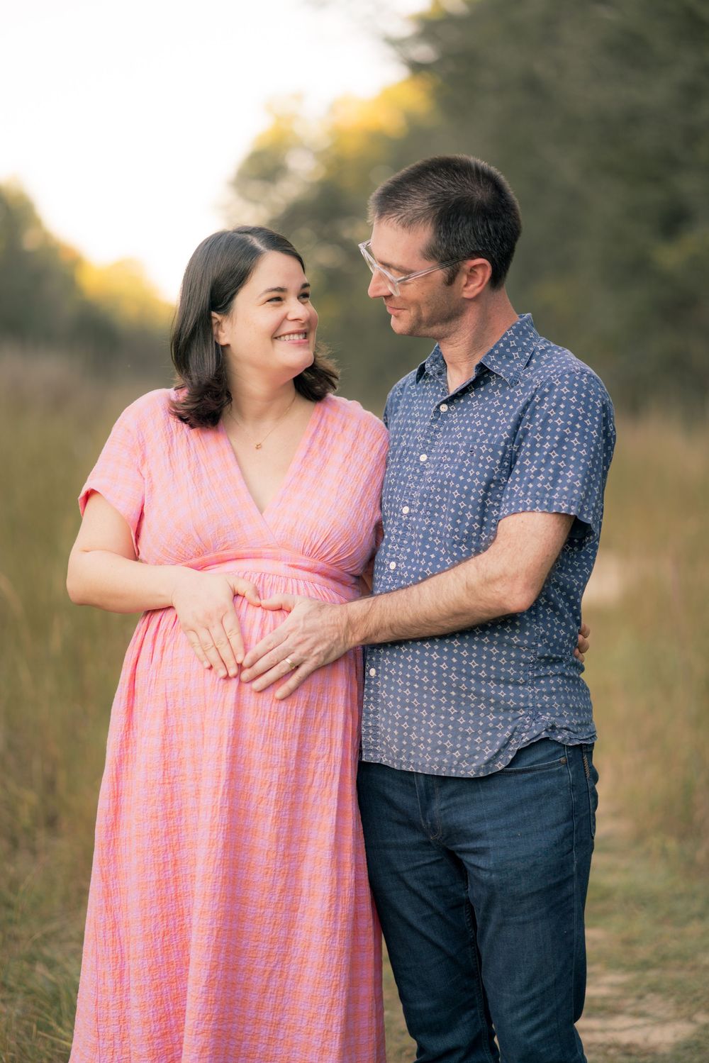 A couple shares an intimate moment outdoors as they celebrate their pregnancy in a pink dress and blue shirt.