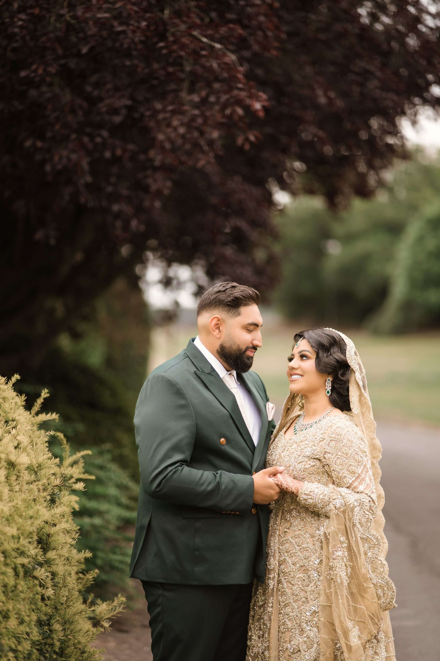 Gorgeous groom and bride staring at each others eyes blissfully, standing in a lushful green garden