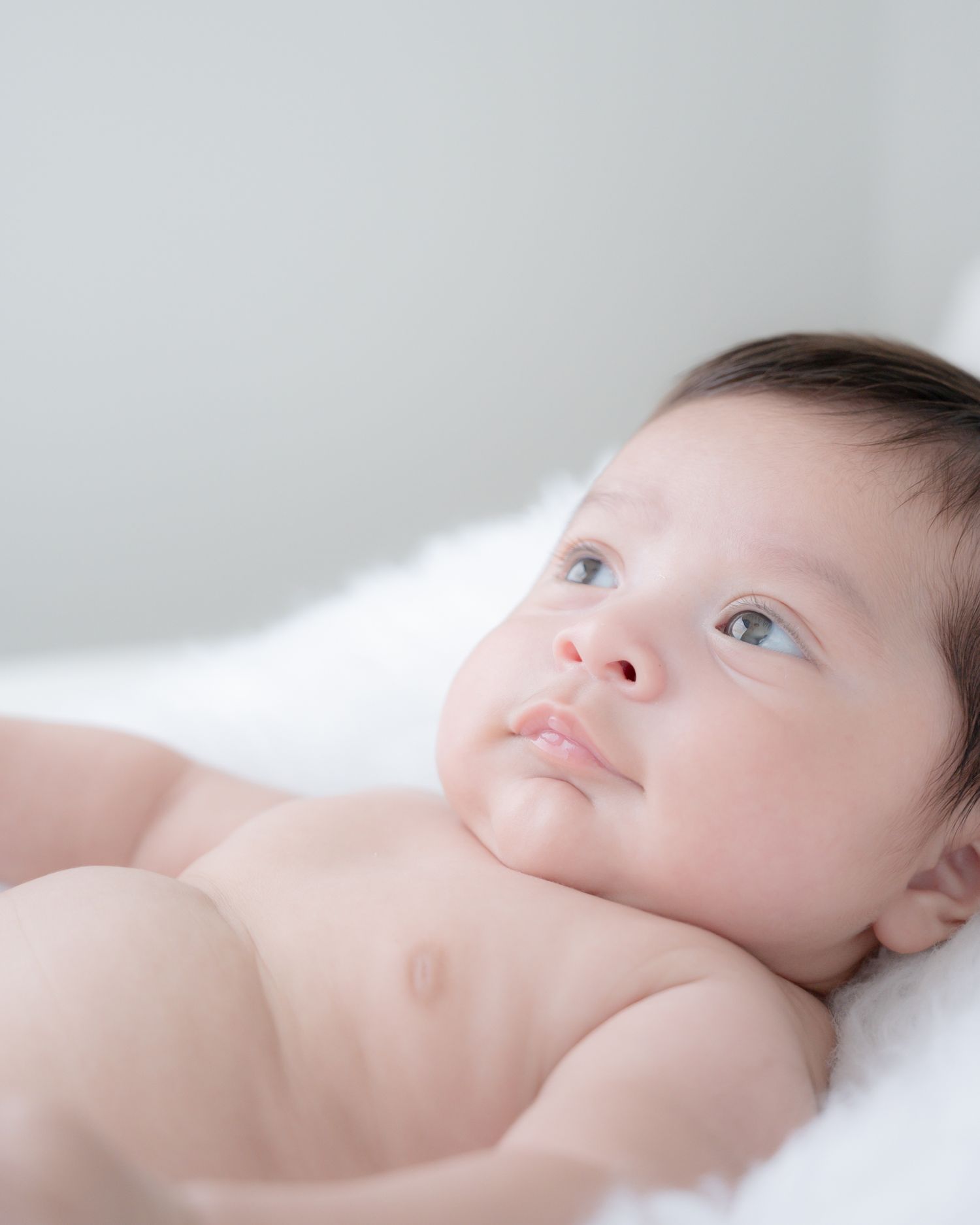 Infant lying on white bedding gazes upward in soft natural light.