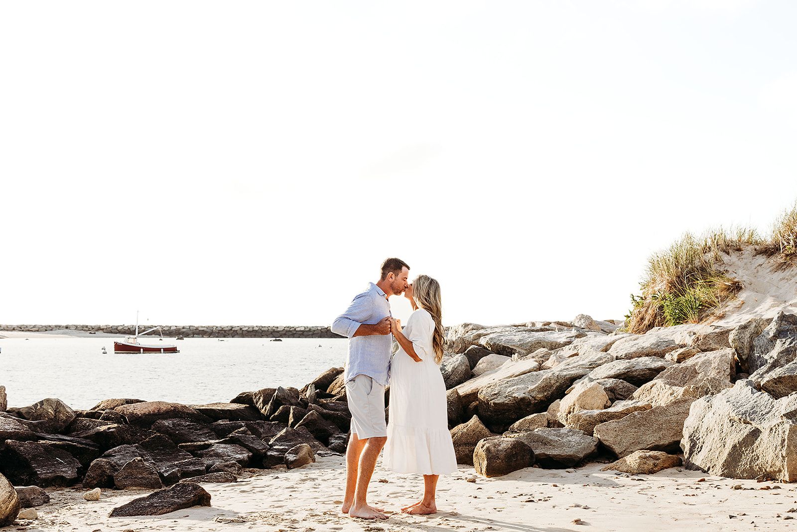 A couple embraces during a romantic beach photoshoot with driftwood and ocean waves in the background.