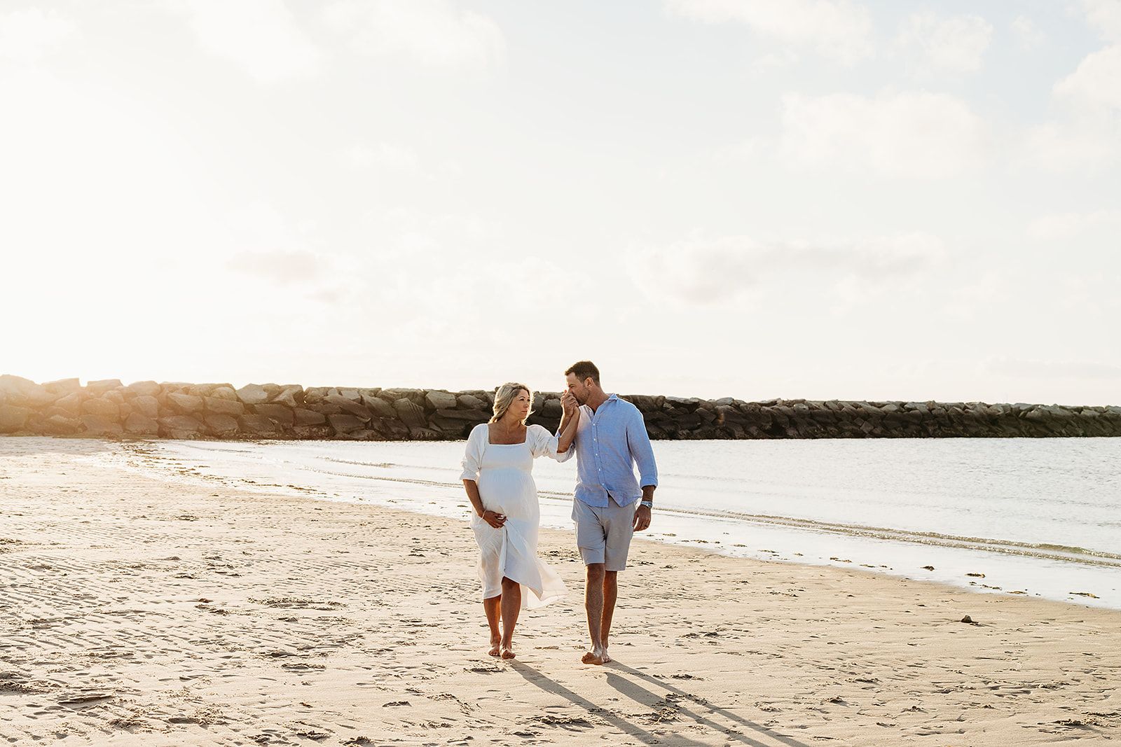 Two people walk hand in hand along a serene beach shoreline at sunset with waves rolling in.