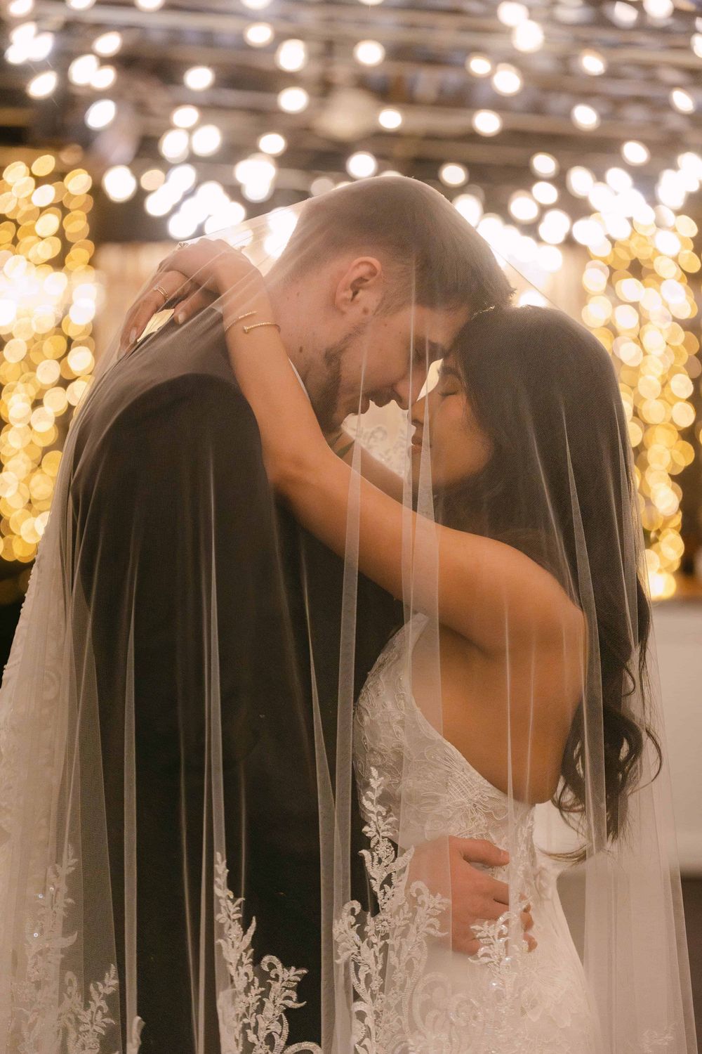 Romantic wedding couple sharing an intimate moment under glowing string lights with beautiful lace veil details.