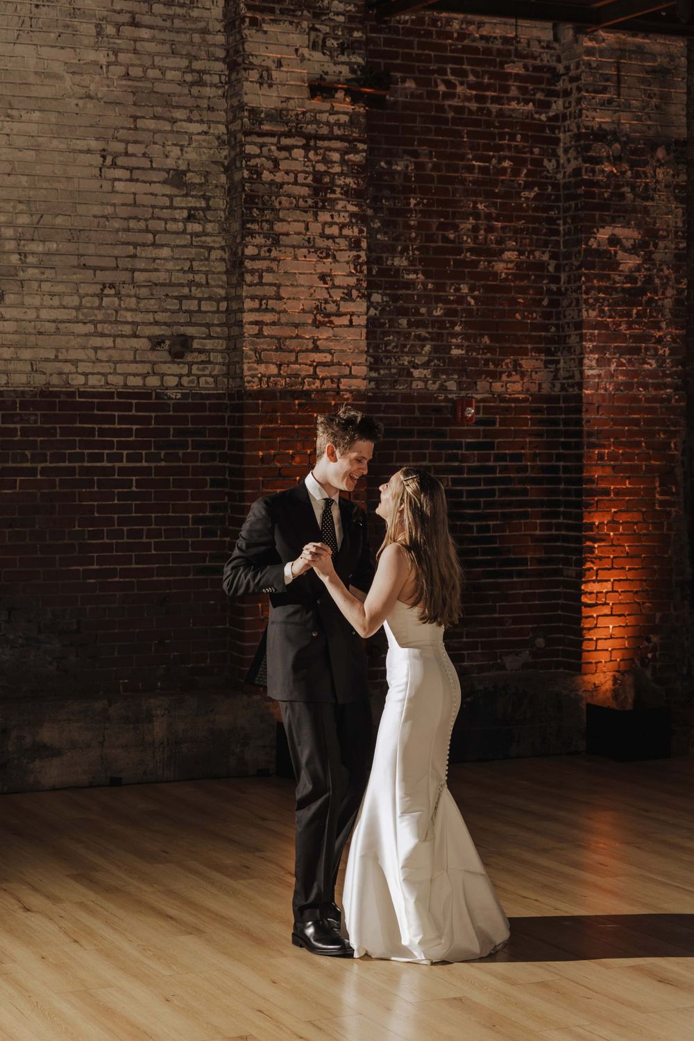 A couple shares their first dance as newlyweds in a rustic brick venue with warm lighting on a wooden floor.
