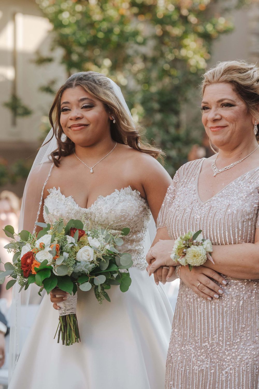 A bride in a white strapless gown stands with companion in beaded dress, both holding elegant floral bouquets outdoors.
