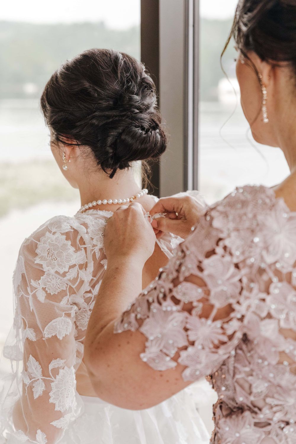 Someone helps fasten a delicate pearl necklace over an intricate lace wedding dress with floral appliques.