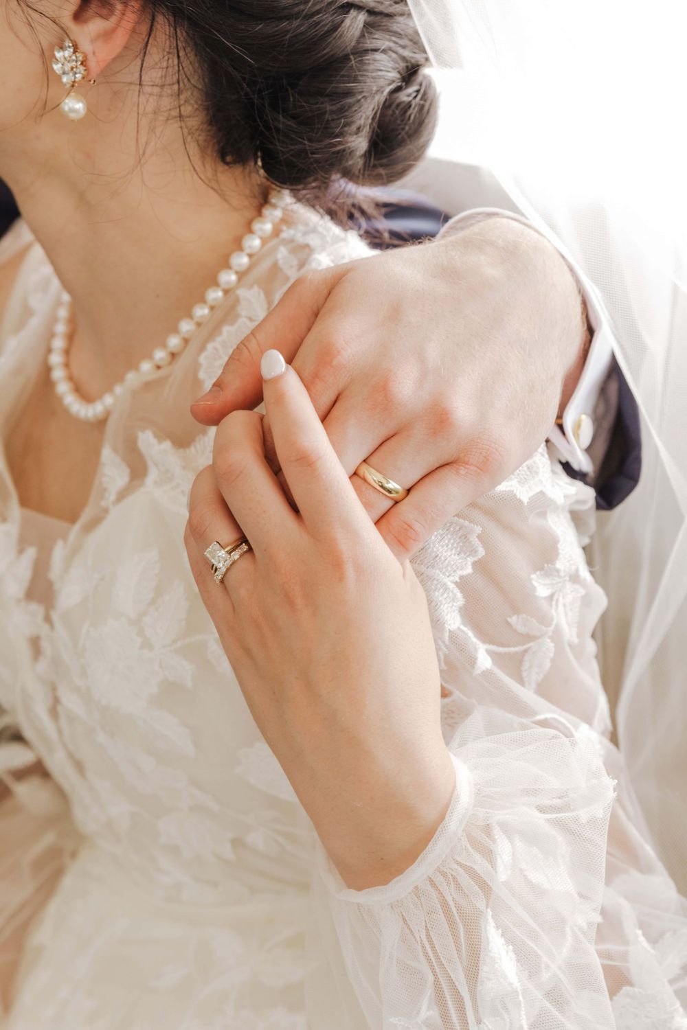 Close-up of elegant bridal jewelry with pearl necklace and diamond rings against white lace wedding dress.