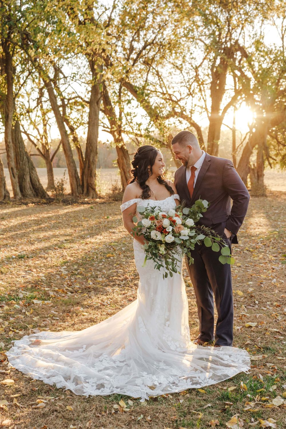 Wedding couple shares an intimate moment at sunset among autumn trees, with the bride holding a lush floral bouquet.