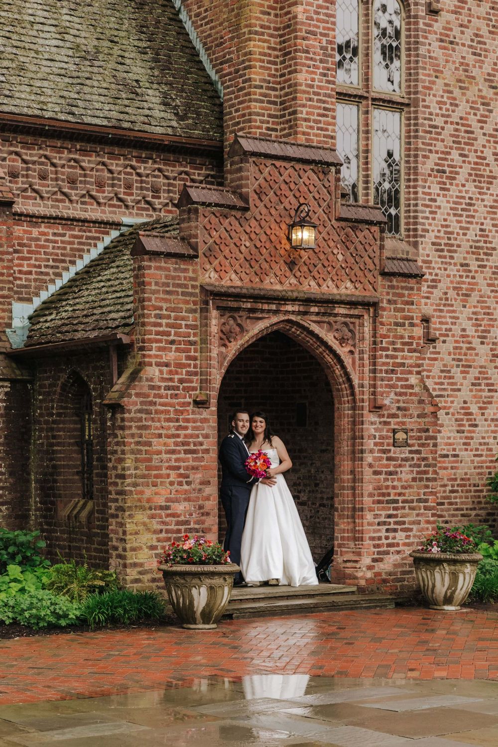 A bride and groom stand together in an ornate brick Gothic church doorway on a rainy day.