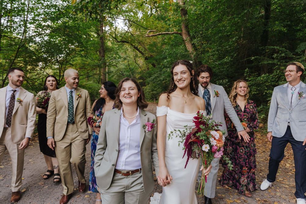 Wedding party walks together on wooded path with bride carrying burgundy floral bouquet in outdoor setting.