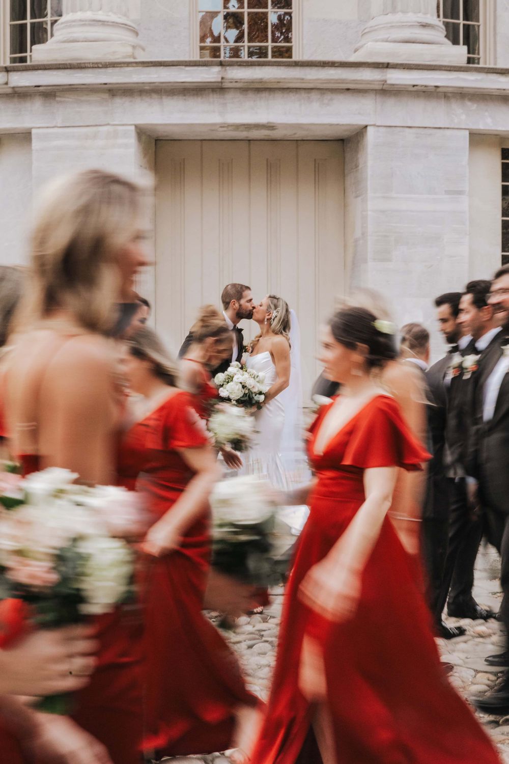 Motion blurred wedding party gathers outside elegant building with bridesmaids in flowing red dresses and white bouquets.