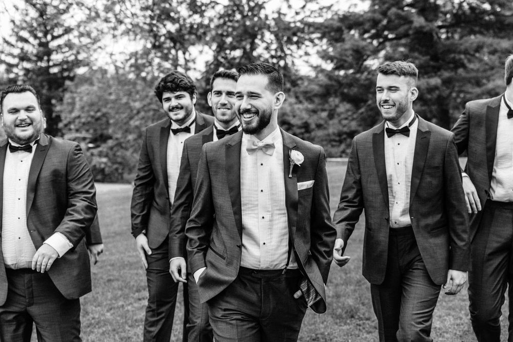 A group of groomsmen in black tuxedos and bow ties laughing while walking together outdoors.