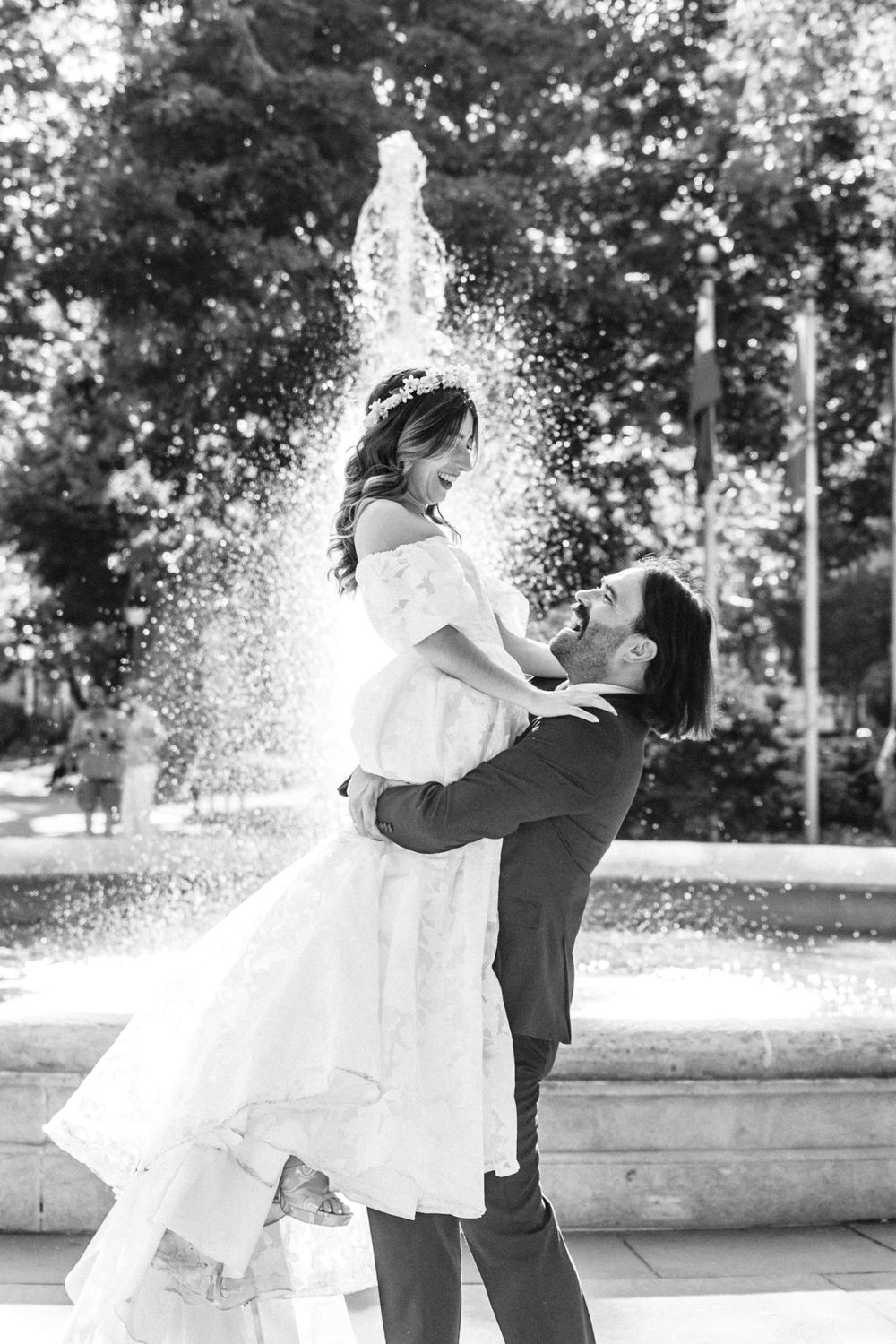 A romantic couple embraces in front of a fountain, with water sparkling behind them in black and white.