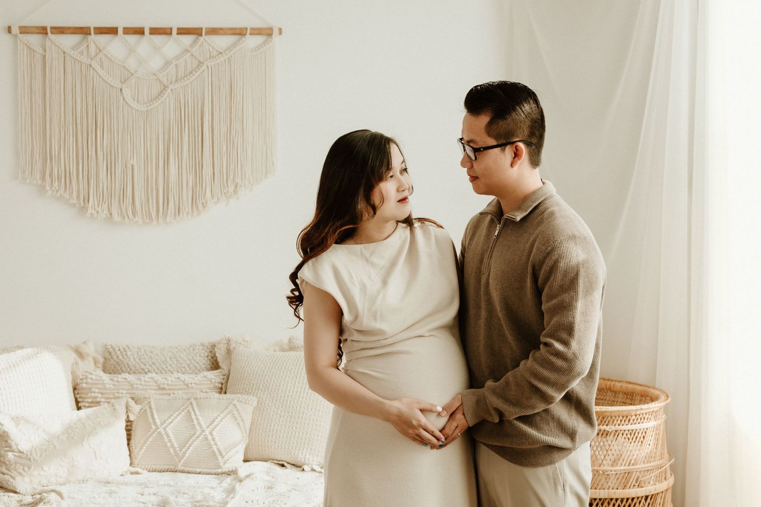 Casual maternity photo with couple in neutral tones standing in cozy room with macrame decor.