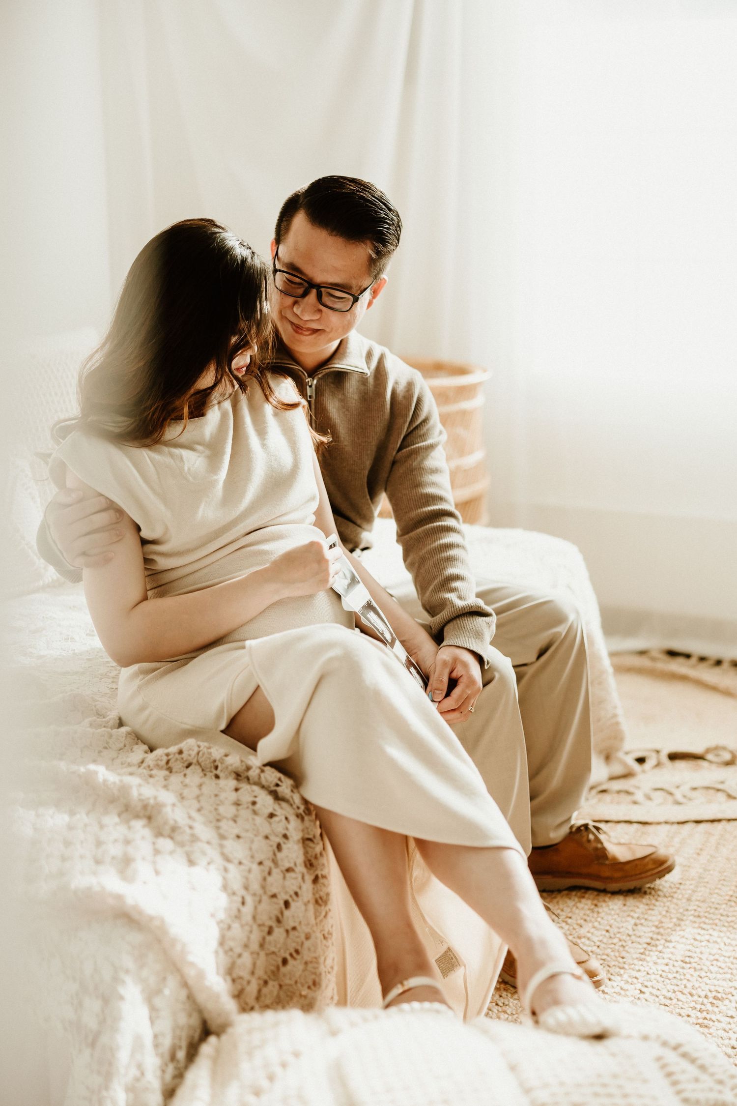 Intimate maternity photoshoot of couple sitting together on couch in light-filled room wearing neutral colors.