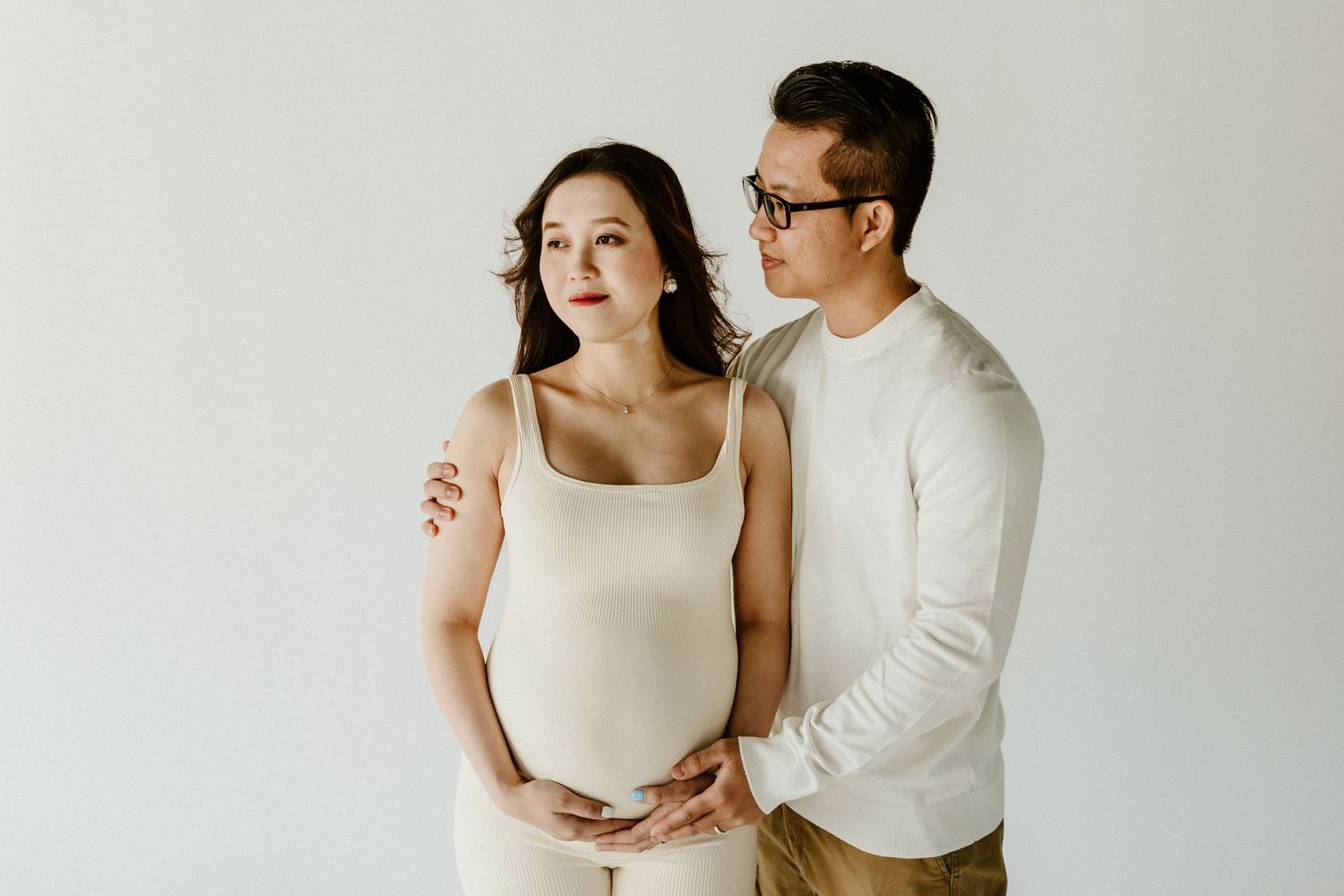 Minimalist maternity photo with expectant couple in white embracing against neutral background.